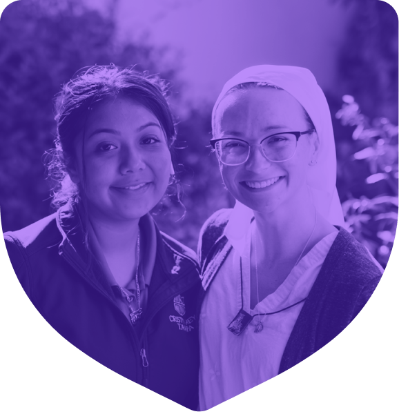 Two women smiling, one wearing glasses, the other in a jacket, stand closely together in an outdoor setting near the Newman Ministry Center.