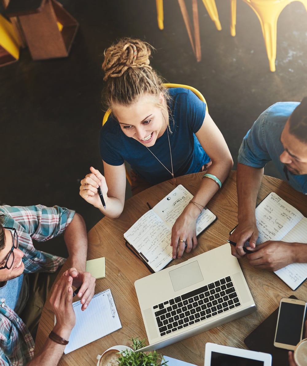 A group of people is collaborating around a table with a laptop, notebooks, and coffee. A woman from a Catholic youth group is smiling and holding a pen, engaging in discussion.