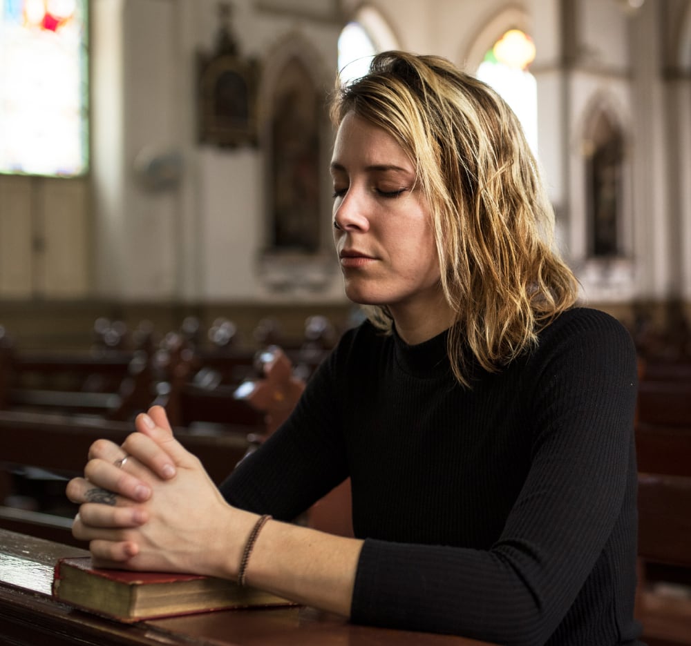 A woman sits in a church pew at the Newman Center, eyes closed and hands clasped over a book, appearing to pray, surrounded by students from nearby Catholic colleges.